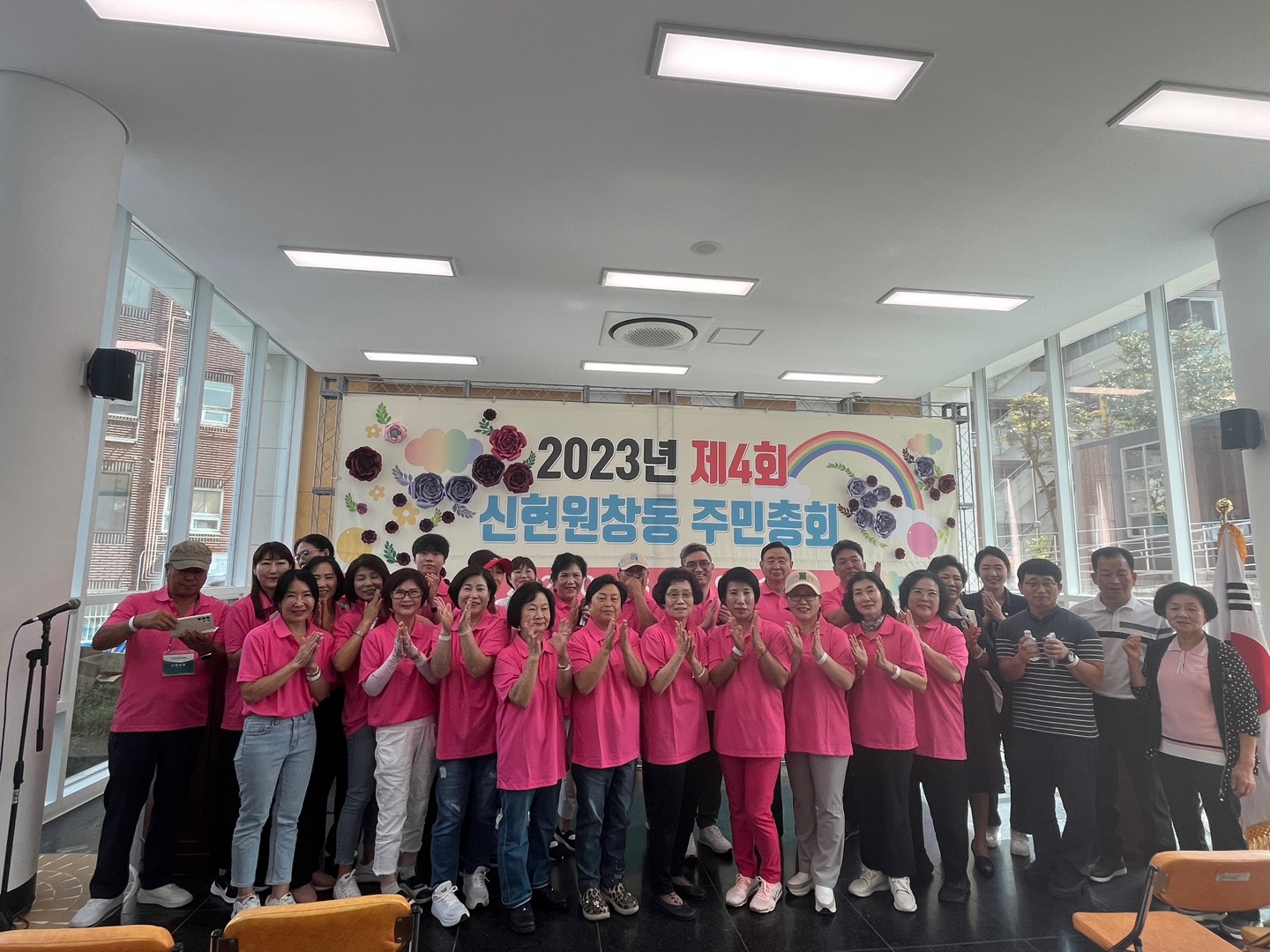 2023년 제 4회 신현원창동 주민총회 현수막 앞에서 참여자들이 분홍색 티셔츠를 맞춰입고 박수를 치며 단체사진을 찍는 현장 모습