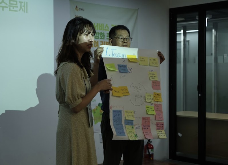 팀1이라는 글과 포스트잇이 붙여진 종이를 든 남성과 여성이 발표하고 있는 모습