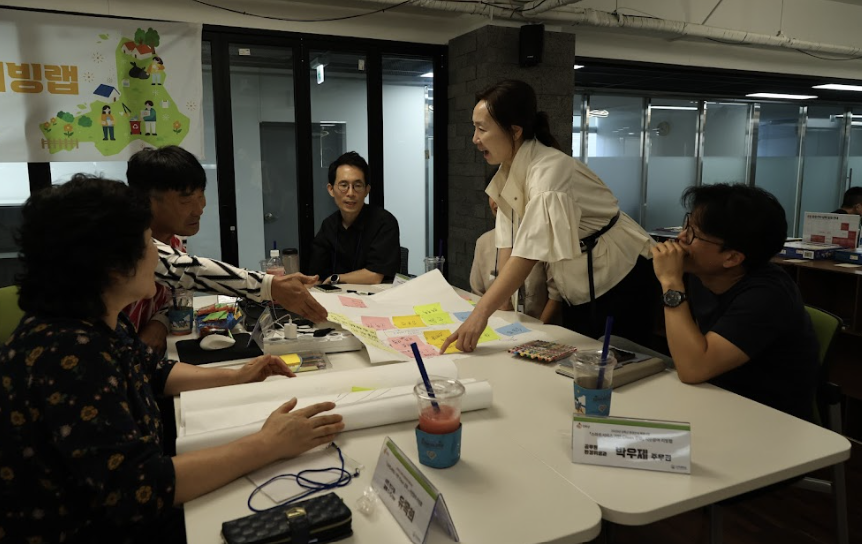 진행자가 참석자들의 테이블에서 포스트를 손가락으로 집으며 설명하고 있는 모습