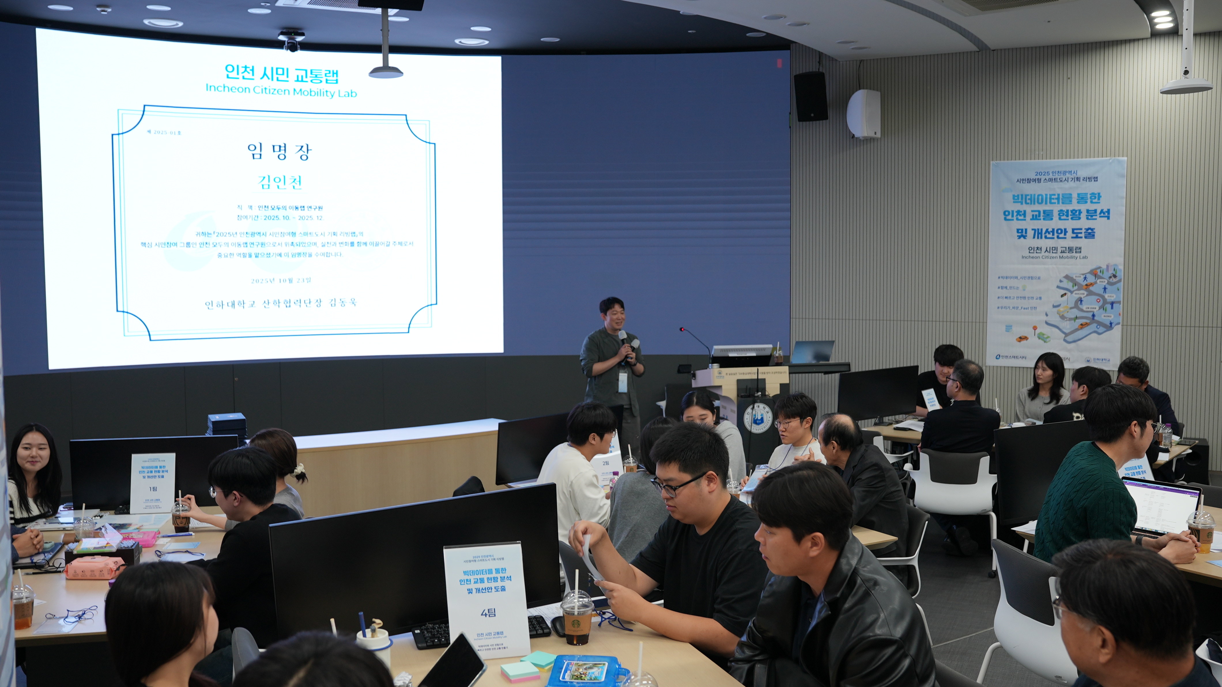임명장이 보이는 프레젠테이션 앞에서 진행자가 설명하고 참석자들은 경청하는 모습